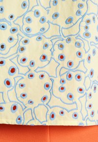 Tissu jaune présentant des motifs circulaires brodés en bleu et rouge, bordés de fil bleu. En dessous, un tissu orange est visible.