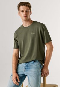 Mladý muž s kučeravými vlasmi, oblečený v olivovo zelenej krátkostičkom tričku a svetlomodrých džínsach, sediaci na drevenej stoličke.