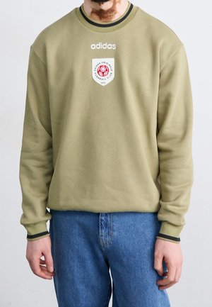 Mann trägt olivgrünes Adidas-Sweatshirt mit rotem Fußball-Logo-Aufnäher, kombiniert mit blauen Jeans, steht vor einem schlichten hellen Hintergrund.