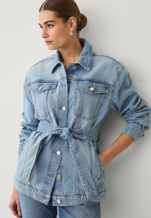 Woman with hair in a bun wearing a belted light blue denim jacket with buttoned chest pockets and rolled-up sleeves.