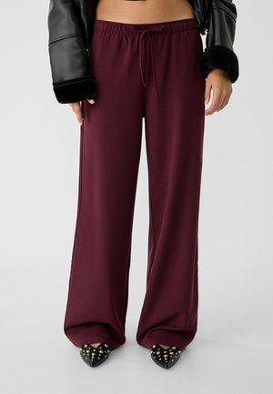 Person trägt weite bordeauxrote Hosen mit Kordelzug in der Taille, schwarze spitze Schuhe mit Nieten und eine schwarze Jacke mit Fellabschlüssen an den Ärmeln.