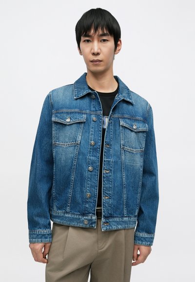 Veste en denim bleu clair avec une patte de boutonnage, deux poches poitrine et un col classique. Présente une texture délavée et des coutures contrastantes.