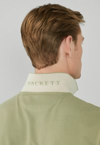 Grön polo med en kontrasterande ljus krage som har texten "HACKETT." Tyget verkar strukturerat, lämpligt för avslappnad användning.