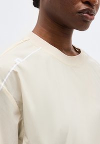 Close-up of a person wearing a beige crewneck shirt with a subtle white line design on the shoulder.