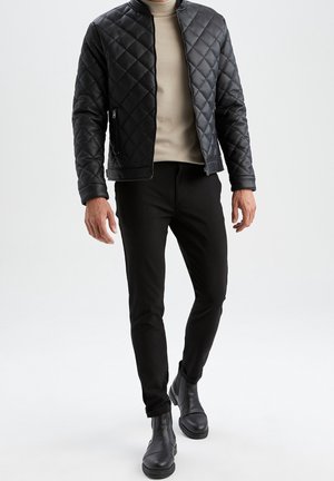 Homme portant une veste matelassée noire, un col roulé beige, un pantalon noir et des bottes en cuir noir marchant sur fond blanc.