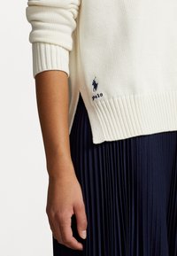 Jersey de punto blanco acanalado con una apertura lateral, que presenta un logotipo bordado en azul oscuro. Combinado con una falda azul marino plisada, destacando las texturas.