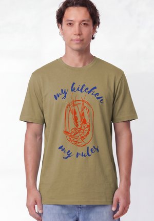 Homme portant un t-shirt vert olive avec un graphisme de crevette rouge et un texte bleu "my kitchen my rules", debout devant un fond uni.