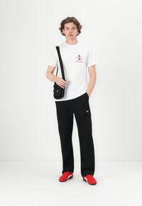 Hvid T-shirt med et lille brunt grafisk motiv, sorte cargo-bukser med lommer og røde sneakers. Skuldertaske spændt over kroppen.