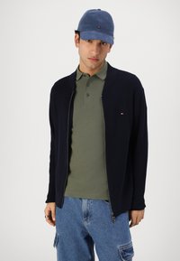 Cardigan navy cu fermoar, purtat peste o cămașă polo verde, asortat cu blugi cargo de un albastru deschis. Casca este de un albastru deschis cu un logo pe partea din față.