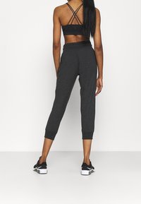 Nainen, jolla on pitkät hiukset, pukeutuneena mustaan nyöritettyyn urheiluliiviin, tummanharmaisiin verkkareihin ja mustiin Nike-tennareihin, seisoo valkoista taustaa vasten.