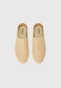 Zapatillas slip-on de lona beige con un upper texturizado, acento de tacón de cuero marrón y suela de goma blanca. Presenta un logo inscrito en la plantilla.