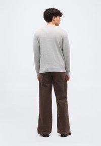 Homme vu de dos portant un pull léger à manches longues gris clair, un pantalon large marron et des chaussures noires sur un fond blanc uni.
