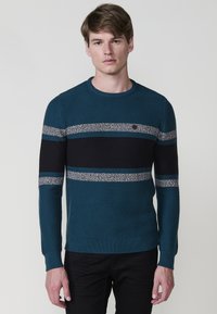 Jersey de punto en verde azulado con rayas horizontales negras y grises, cuello redondo y puños acanalados. Presenta un pequeño logotipo en el pecho.