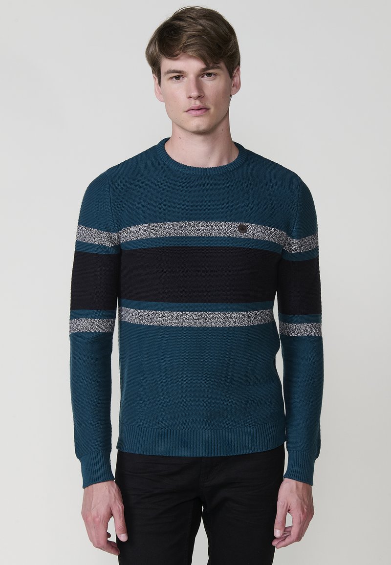 Jersey de punto en verde azulado con rayas horizontales negras y grises, cuello redondo y puños acanalados. Presenta un pequeño logotipo en el pecho.