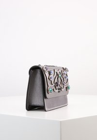 Pieni hopeanvärinen käsilaukku, jossa on koristeellisia monivärisiä jalokiviä ja monimutkaisia kuvioita, asetettuna valkoiselle pinnalle yksinkertaisen taustan edessä.