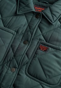 Chaqueta acolchada verde oscuro con cuello, que presenta botones de presión negros, dos bolsillos en el pecho y un parche de marca en el bolsillo izquierdo.