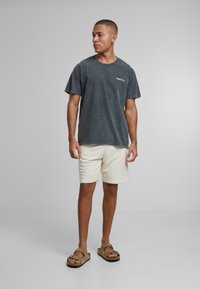 Tmavohnedé tričko s krátkym rukávom a jemným logom, spárované s krémovými šortkami a hnedými sandálmi, na neutrálne pozadie.