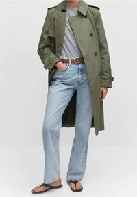 Olivgrüner Trenchcoat, graues T-Shirt, hellblaue Jeans mit geradem Bein, brauner Gürtel und schwarze Flip-Flops. Glatter Stoff und lässiger Stil.