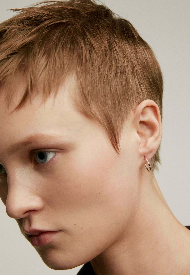Aro de plata llevado en un cabello corto y texturizado. El primer plano muestra la forma redonda del aro y su acabado de metal liso, resaltando la simplicidad.