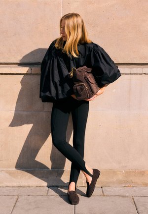 Frau mit blonden Haaren, die ein schwarzes voluminöses Oberteil, schwarze Leggings und braune Loafers trägt und einen braunen Leder-Rucksack an eine Steinmauer hält.