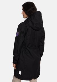 Juodas vandeniui atsparus džemperis su kapišonu, prisiūtu liemeniu ir purpuriniu logotipu ant kairiojo rankovės. Yra baltos atspindinčios juostelės apatinėje dalyje.