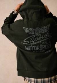 Tmavozelená mikina s kapucňou s veľkým textúrovaným grafickým motívom na chrbte, ktorý zobrazuje krídla a nápis. Pružné manžety a pás.