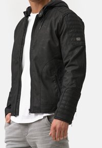 Mustat nahkatakki hupparilla, jossa on tikkaukset hartioissa, vetoketjukiinnitys ja sivutaskut. Sileä pinta ja muotoiltu malli.