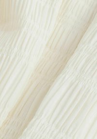 Flødefarvet stof med et struktureret, opsamlet design med lodrette folder over det hele, der viser et glat og let materiale.