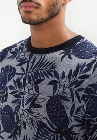 Nahaufnahme des unteren Gesichts und des Halses eines Mannes, der einen grauen Pullover mit dunkelblauen tropischen Blatt- und Ananasmustern trägt.