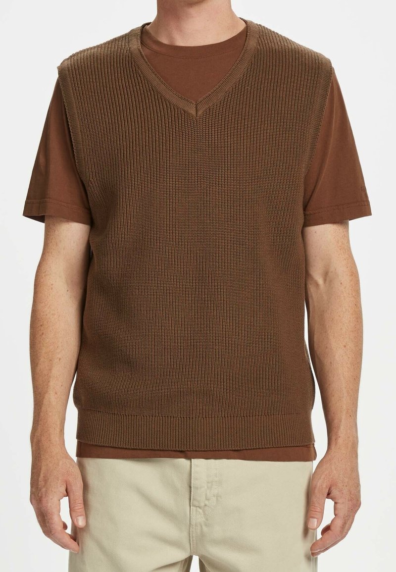 Homme portant un pull sans manches en maille marron à col en V par-dessus un t-shirt marron à manches courtes avec un pantalon beige sur un fond uni.