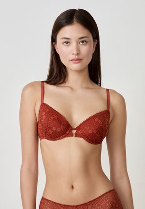 Reggiseno in pizzo rosso con coppe imbottite, spalline regolabili e bordi ondulati. Presenta motivi floreali e un piccolo dettaglio a taglio centrale.