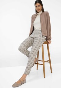 Hellbraune Bomberjacke über einem cremefarbenen Rollkragenpullover, kombiniert mit gemusterten beigen Hosen mit einem abstrakten Design. Graue Ballerinas runden das Outfit ab.
