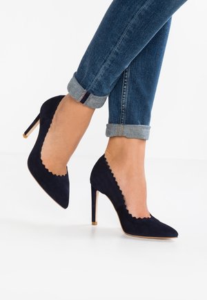 Chaussures à talons hauts en cuir suédé bleu marine avec des bords festonnés, des bouts pointus et un talon stiletto fin, accompagnées de jeans en denim retroussés.