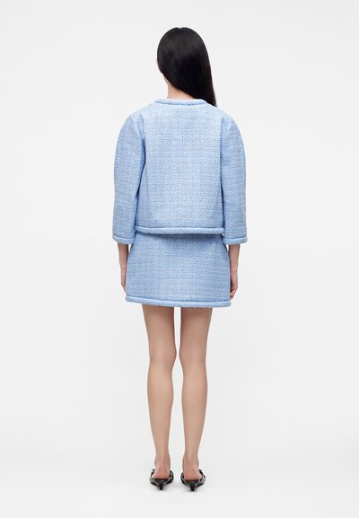 Femme aux longs cheveux foncés portant une veste texturée bleu clair et une jupe assortie, debout de dos et chaussée de talons noirs.
