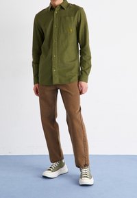 Camisa verde oliva de manga larga con bolsillo frontal y logo, combinada con pantalones marrones y zapatillas verdes, sobre un fondo liso.