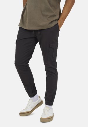Uomo che indossa pantaloni jogger cargo neri, calzini bianchi e scarpe da ginnastica bianche con accenti beige, in piedi con le mani nelle tasche.