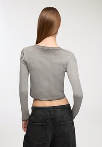 Szary krótki top z długim rękawem wykonany z gładkiego materiału, z okrągłym dekoltem i elastyczną teksturą. Noszony z czarnymi dżinsami z wysokim stanem.