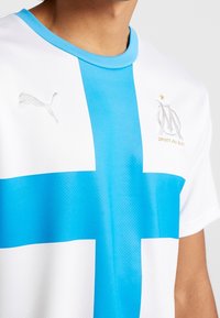 Valkoinen urheilupaita, jossa on sininen ristikkokuvio, sininen kaulus ja hopea Puma-logo. Marseille-embleemi näkyy vasemmalla puolella.