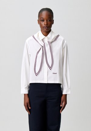 Femme portant une chemise blanche à boutons avec une écharpe rayée nouée autour du cou et un pantalon foncé, debout devant un fond uni.