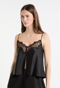 Zwarte satijnen camisole met kanten afwerking bij de halslijn en knopen aan de voorkant. Voorzien van fijne verstelbare bandjes en een uitlopende onderkant.