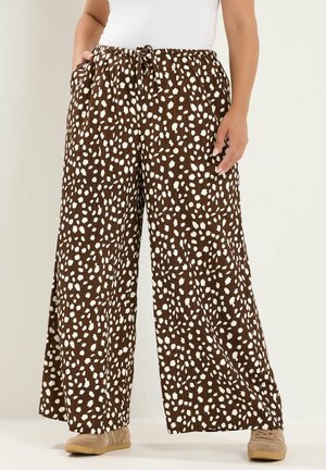 Pantalones marrones de pierna ancha con manchas blancas irregulares, cintura elástica con cordón, usados con zapatos beige y una camiseta blanca.