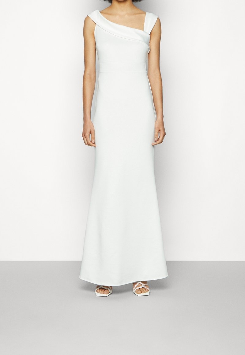 Femme portant une robe blanche longue ajustée avec un décolleté asymétrique et des sandales blanches à lanières, debout devant un fond uni.