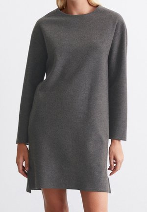 Kobieta ubrana w szarą, długorękawową, dzianinową sukienkę do kolan z bocznymi rozcięciami, stojąca na tle białej ściany.