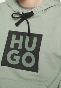 Světle zelená mikina s černým obdélníkovým nášivkem s výraznými zelenými písmeny vyjadřujícími "HUGO." Obsahuje černou šňůrku u límce.