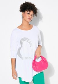 Weißes Langarmhemd mit silbernem Herzmuster, kombiniert mit grünen Hosen und einer pinken Handtasche. Glattes Material und lockere Passform.
