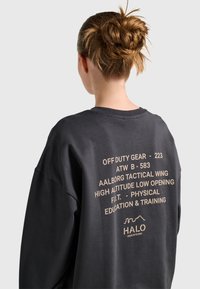 Vrouw met een rommelige knot die een donker sweatshirt draagt met beige tactische stijl tekst op de achterkant over hoogtetraining en onderwijs.