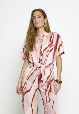 Vrouw met lang haar, gekleed in een roze en bruine gestreepte kortarmige knoopblouse en bijpassende high-waist broek tegen een effen achtergrond.