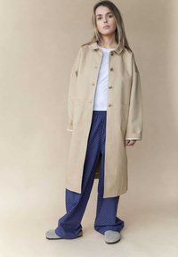 Beige langer Mantel mit braunen Knöpfen, kombiniert mit navyblauen Polka-Dot-Hosen und grauen Hausschuhen. Glattes Material und lässiger Schnitt.