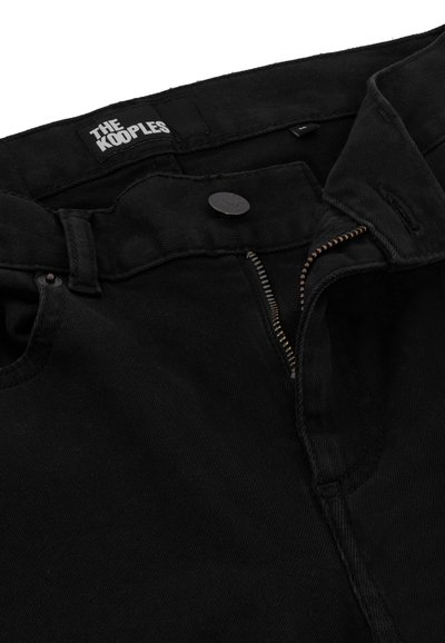 Jeans de mezclilla negra con cremallera, herrajes de metal y costuras visibles. La cinturilla presenta una etiqueta de logo. Textura suave.