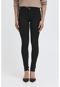 Jean skinny noirs avec un design classique à cinq poches, fermeture à bouton et un tissu lisse, légèrement texturé. Assortis avec des talons marron foncé.